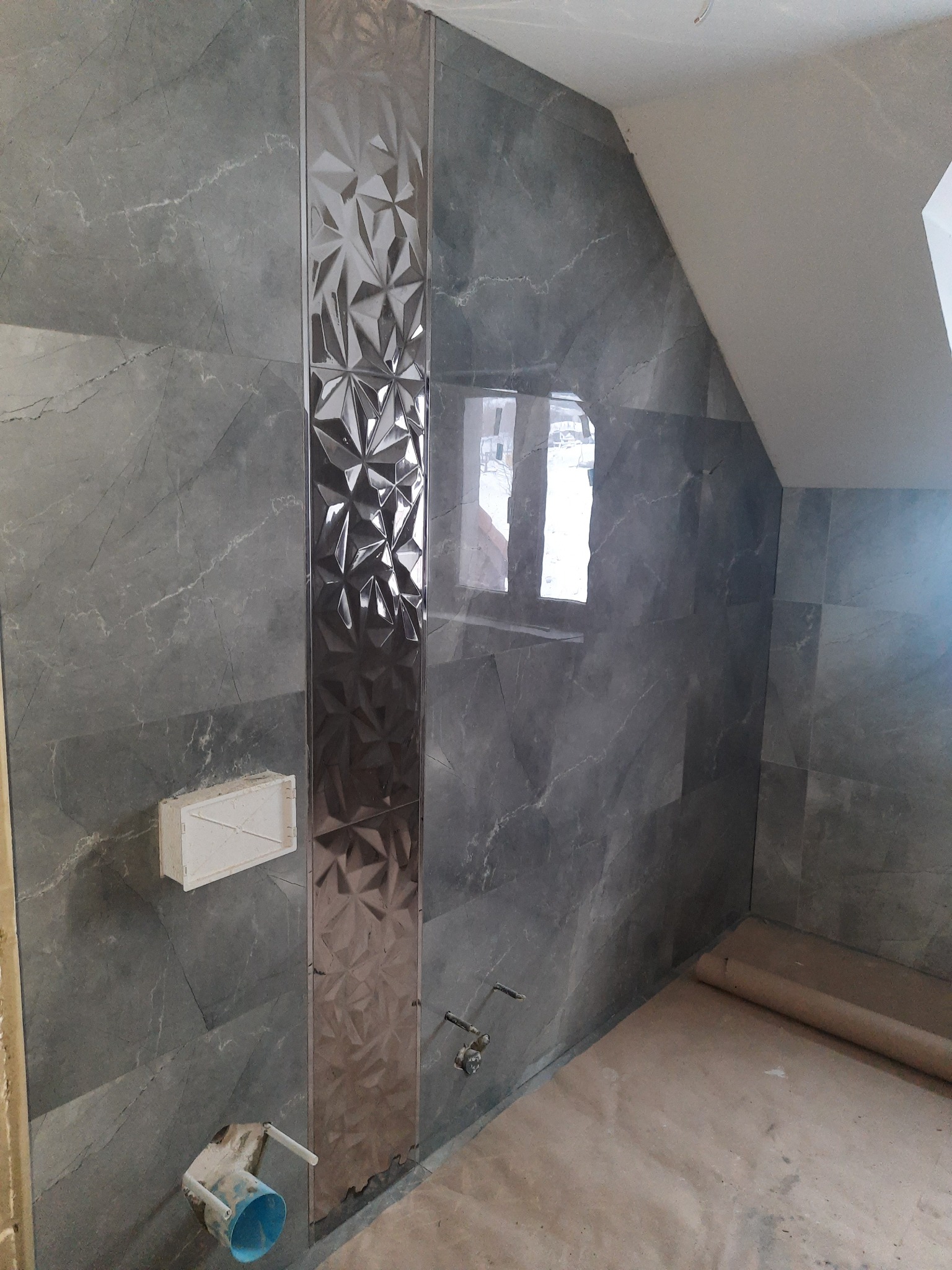 Ściana w łazience wyłożona szarymi płytkami imitującymi marmur, z dekoracyjnym pionowym pasem płytek o trójwymiarowej strukturze, widoczne elementy instalacji wodnej i przygotowane podłoże.