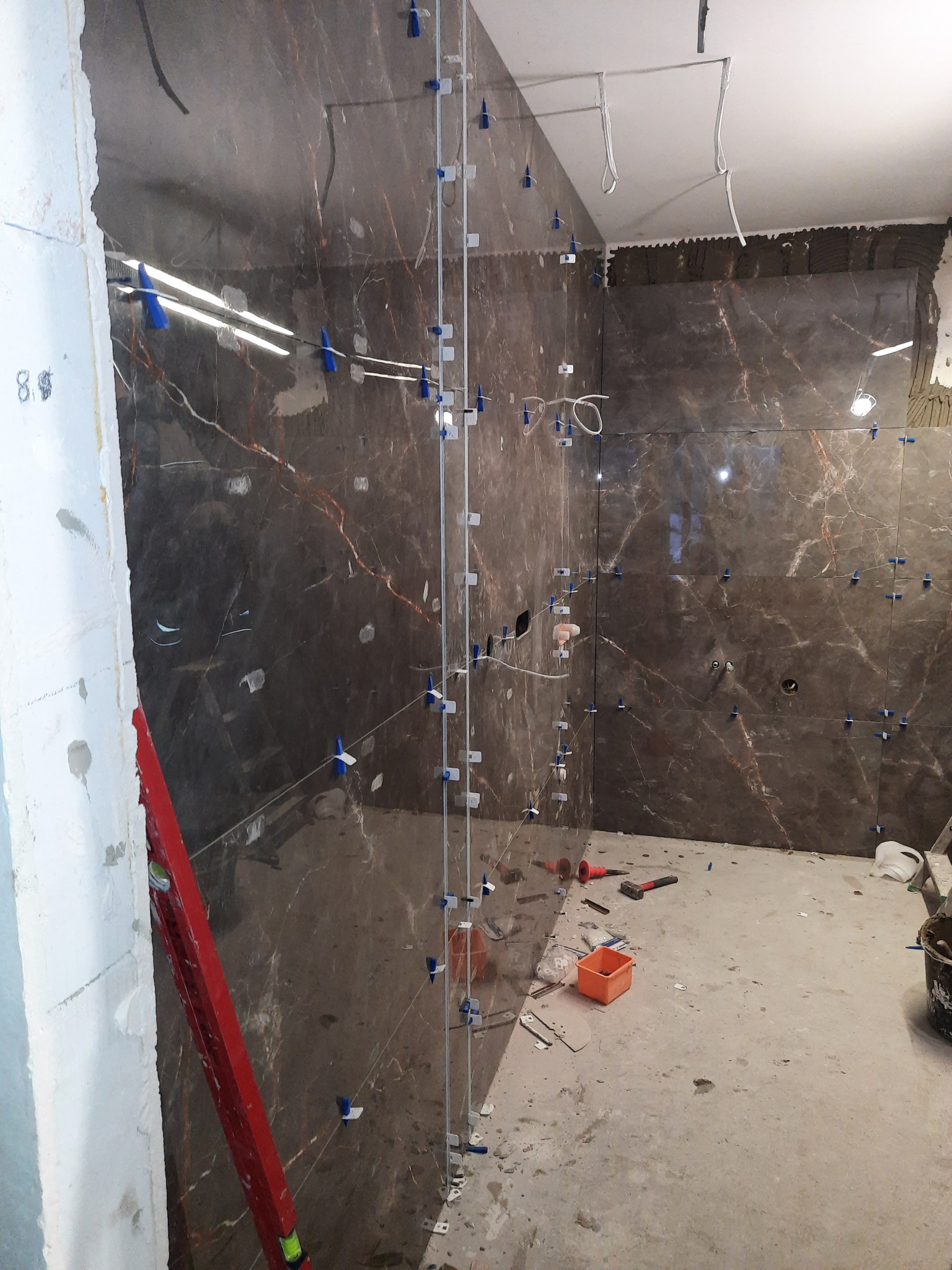 Montaż płytek imitujących marmur na ścianie łazienki z użyciem systemu poziomowania płytek, widoczne narzędzia i poziomica.