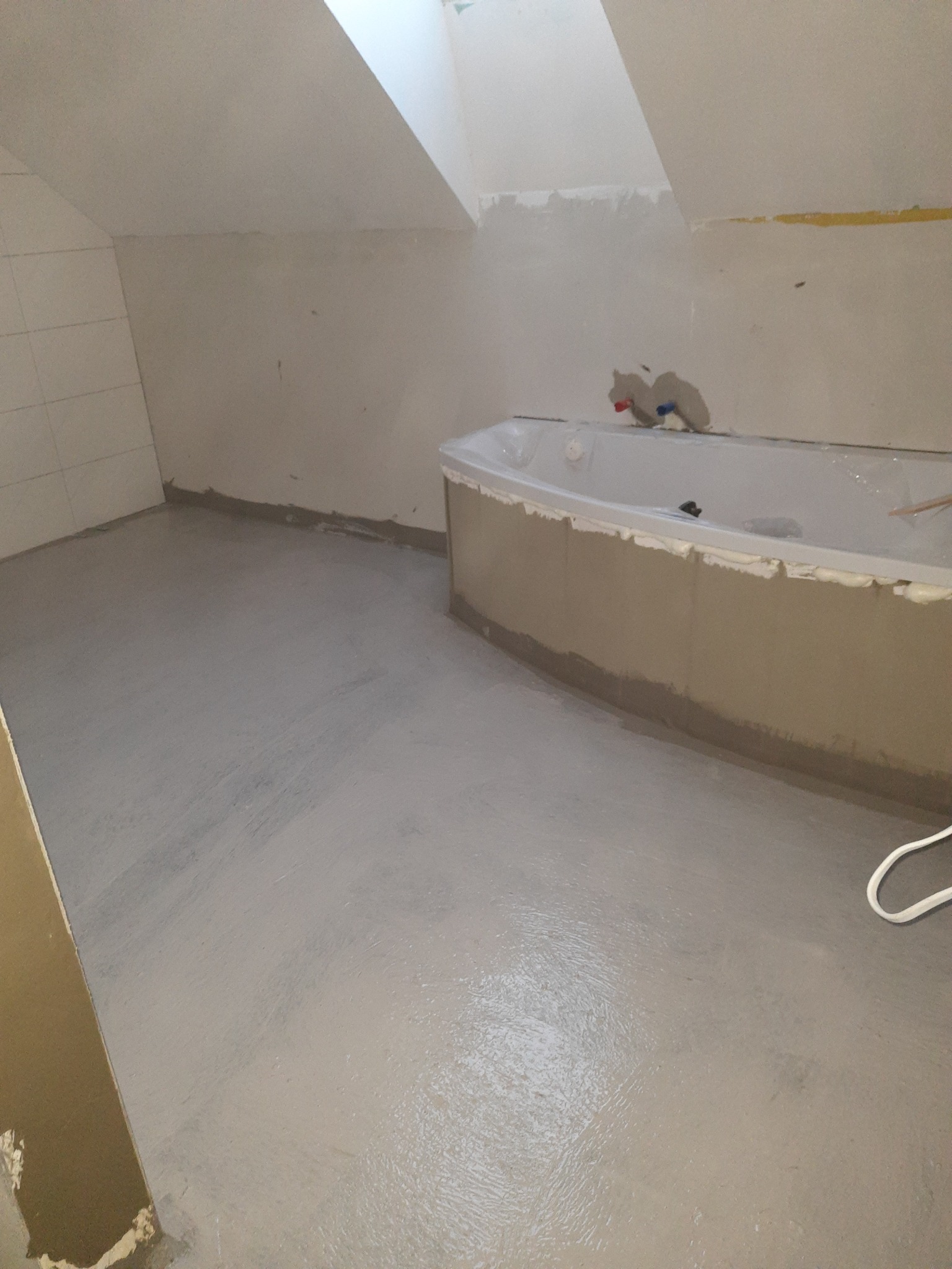 Łazienka w trakcie prac wykończeniowych w poddaszu: widoczna wanna obłożona pianką montażową i świeżo wylana, jasnoszara wylewka na podłodze, płytki częściowo położone na ścianie.