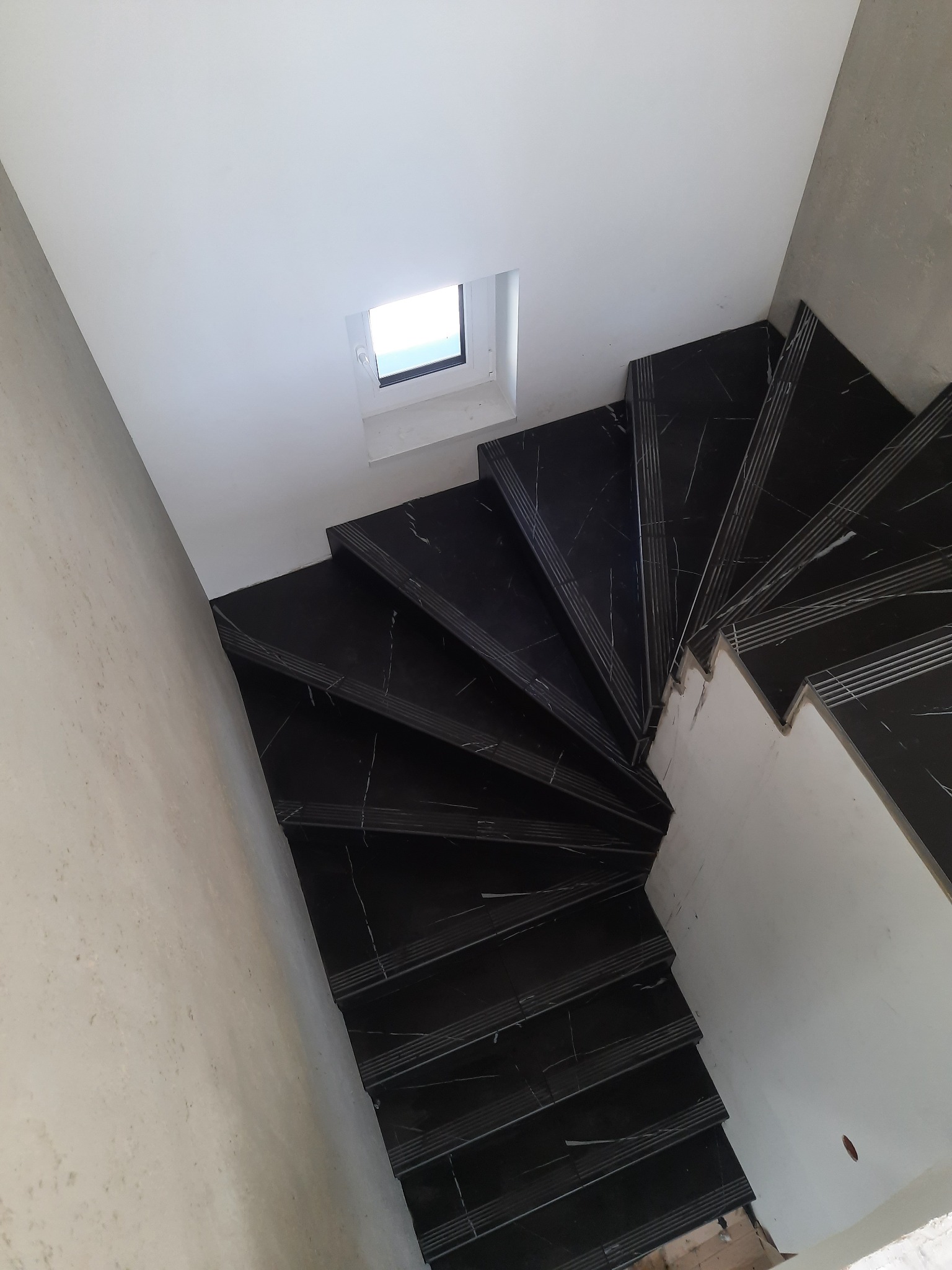 Widok z góry na spiralne schody wyłożone czarnymi płytkami z białym żyłkowaniem, widoczne surowe ściany i małe okno dachowe.