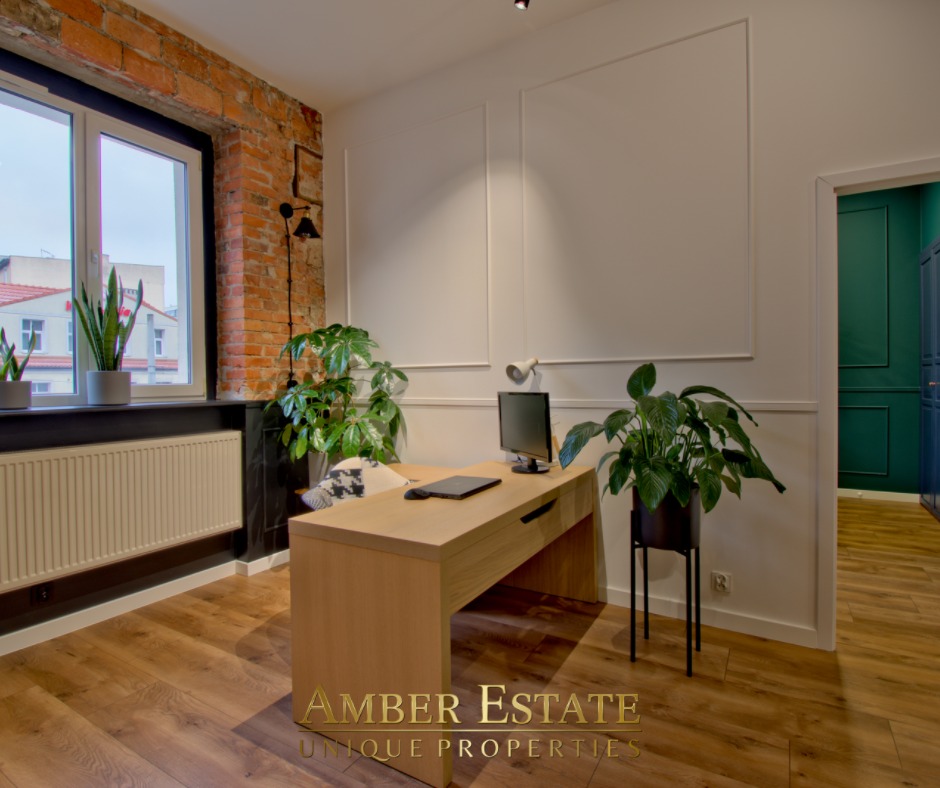 Jasne biuro w mieszkaniu z ceglaną ścianą, drewnianym biurkiem, zielonymi roślinami w doniczkach i widokiem na miasto z okna.