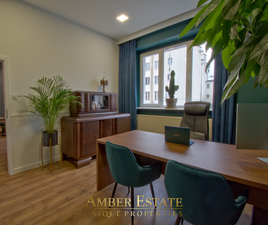 Nowoczesne biuro z drewnianym biurkiem, zielonymi krzesłami i roślinami doniczkowymi, w tle okno z widokiem na budynek mieszkalny.