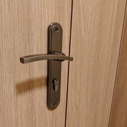 DoorPRO Tomasz Kulesza - Zamknięte drzwi wewnętrzne z drewnianą okleiną i metalową klamką w odcieniach brązu. Widoczny zamek. Ujęcie z bliska, skupione na detalu.