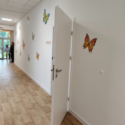 DoorPRO Tomasz Kulesza - Otwarta biała stolarka drzwiowa z srebrną klamką w korytarzu z dekoracjami motyli na ścianach i panelami podłogowymi. Widoczne osoby w tle.