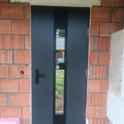 DoorPRO Tomasz Kulesza - Grafitowe drzwi zewnętrzne z szybą, osadzone w surowym murze z czerwonej cegły. Widoczne przewody elektryczne i fragment samochodu w szybie.