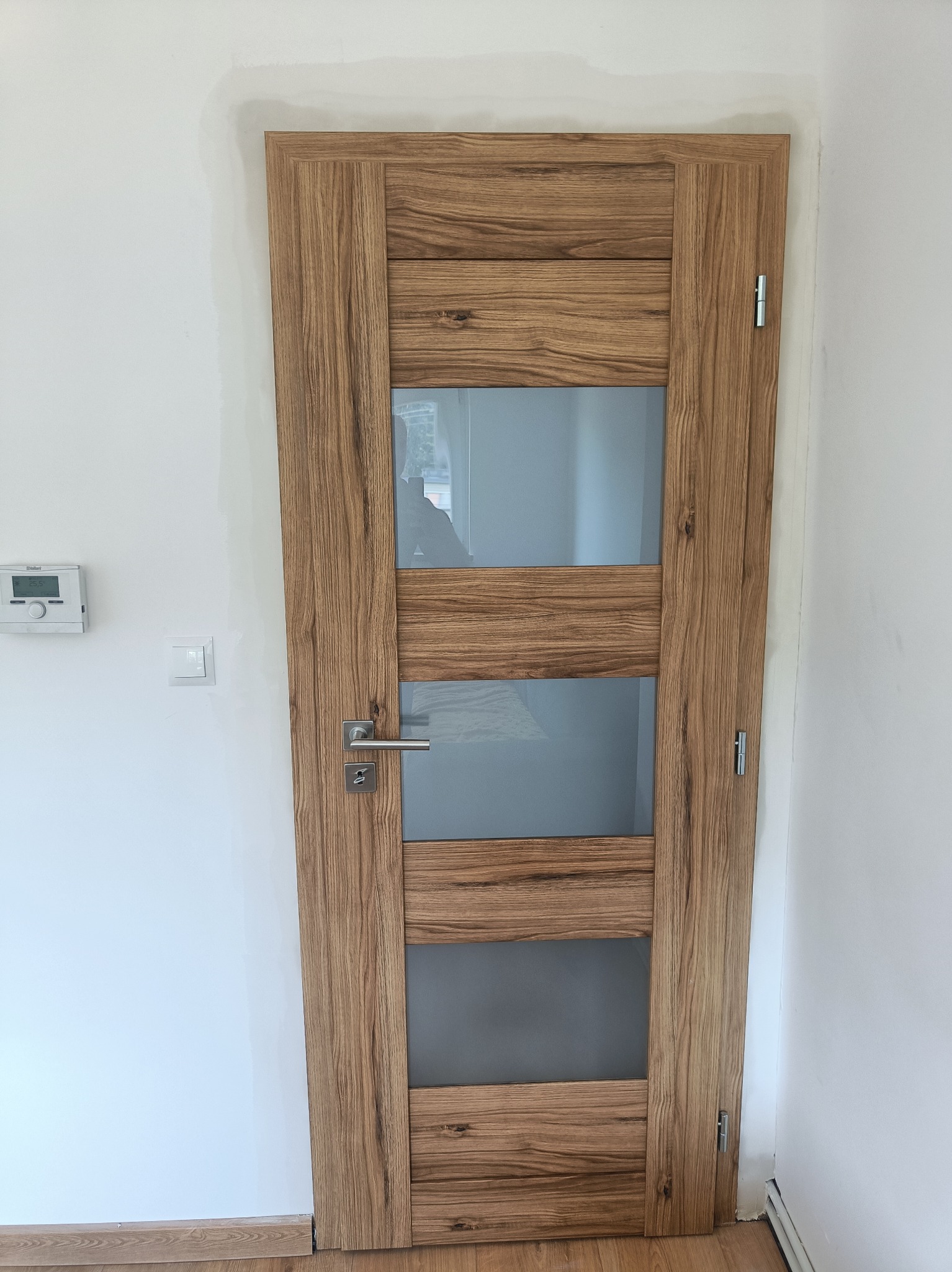 Dębowa okleina na skrzydle drzwiowym z trzema szklanymi, matowymi prostokątami; widoczny srebrny klucz w zamku i biała ściana z termostatem.