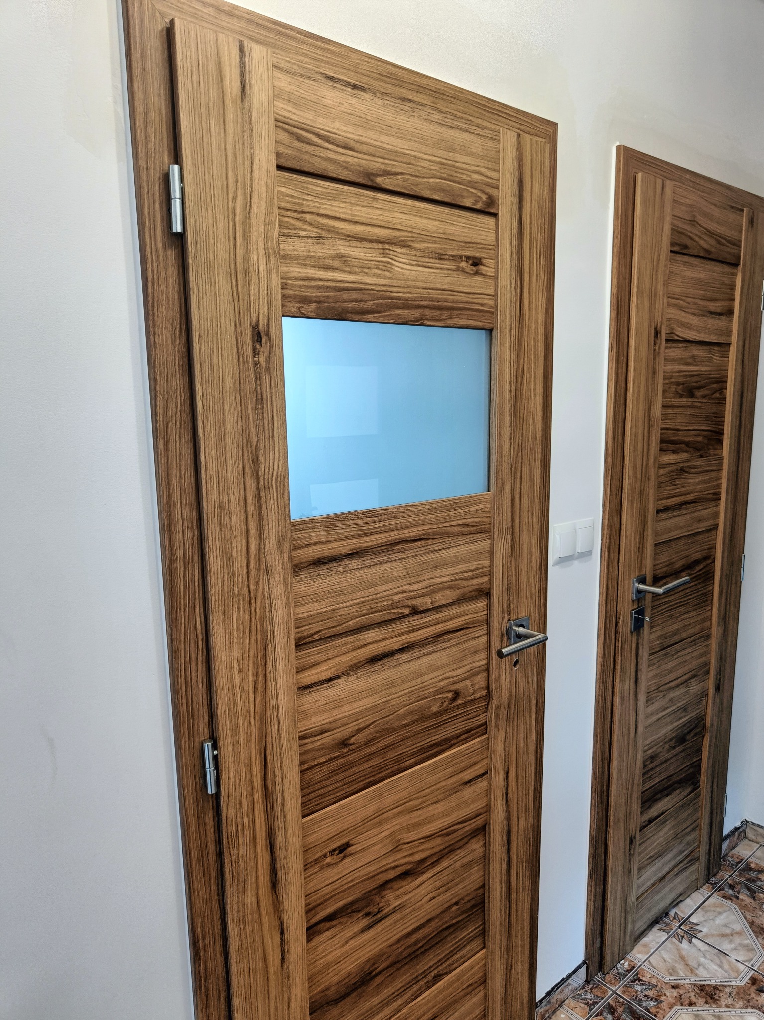 Dwie dębowe drzwi wewnętrzne z widocznym usłojeniem, jedne z matową szybą, zamontowane w białej ścianie z widocznym przełącznikiem światła.