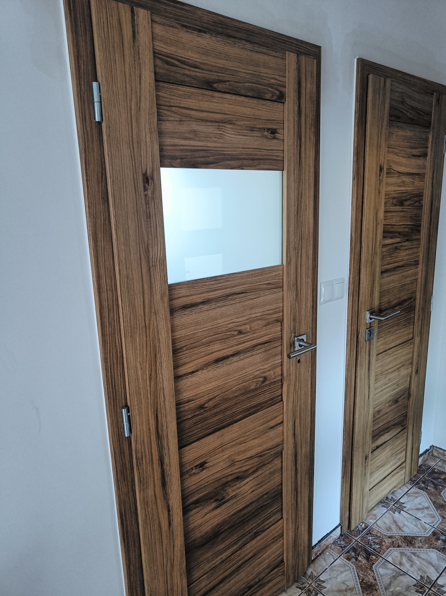 Dwie drewniane drzwi wewnętrzne z wyraźnym usłojeniem, jedne z prostokątną szybą, zamontowane w białej ścianie, widoczne zawiasy i klamki.