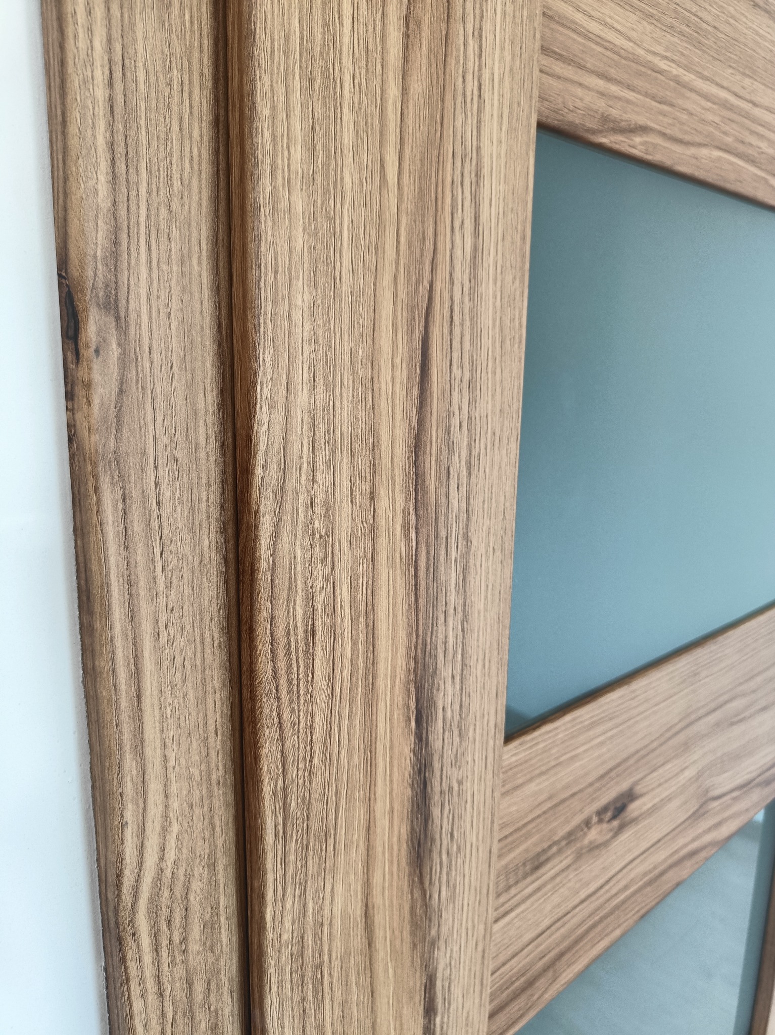 Fragment drewnianej ramy drzwi z matową szybą w odcieniu błękitu, widoczne usłojenie drewna i biała ściana w tle.