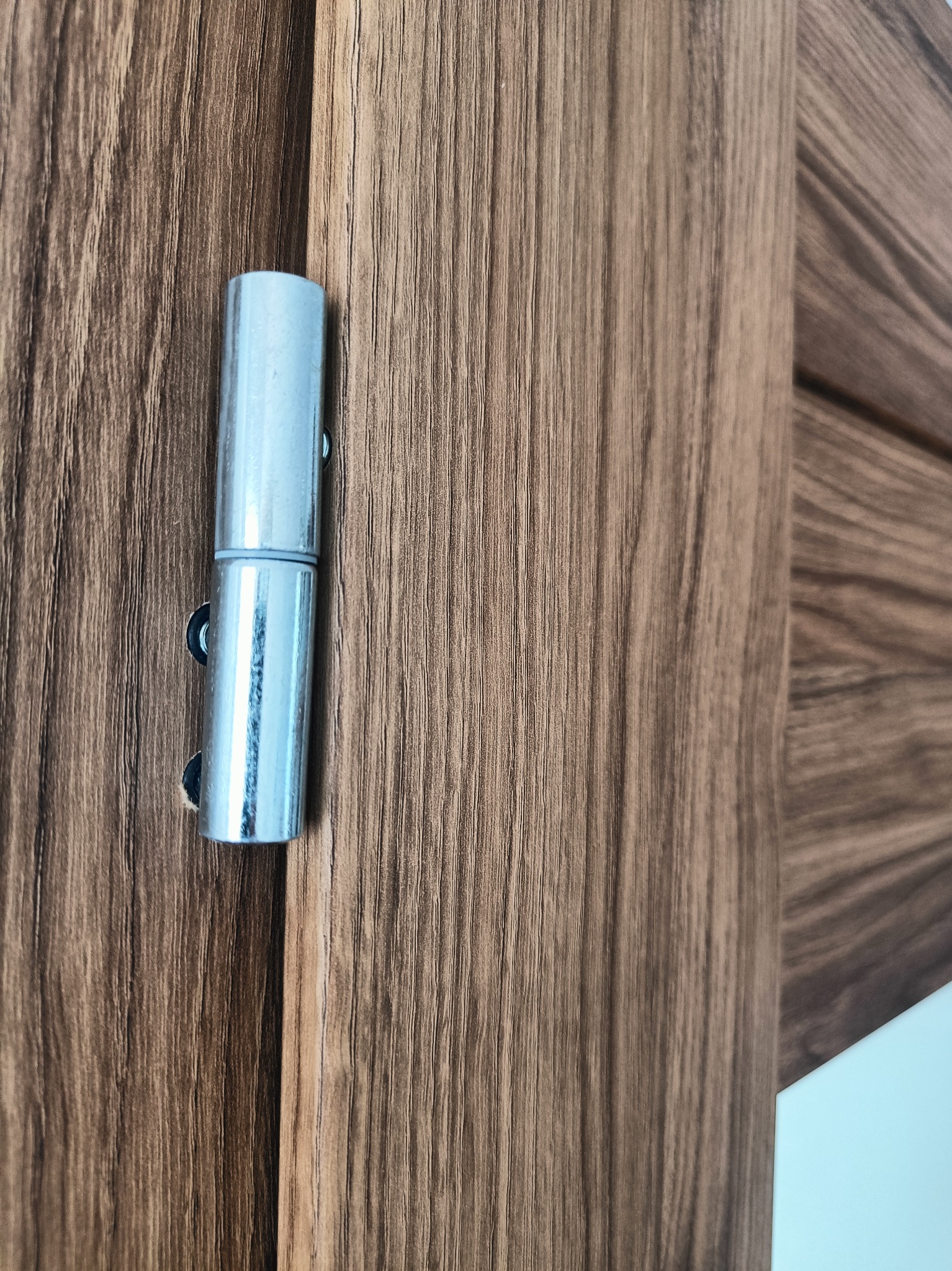 Szczegół chromowanego zawiasu na brązowych, drewnopodobnych drzwiach wewnętrznych z widocznym wzorem usłojenia.