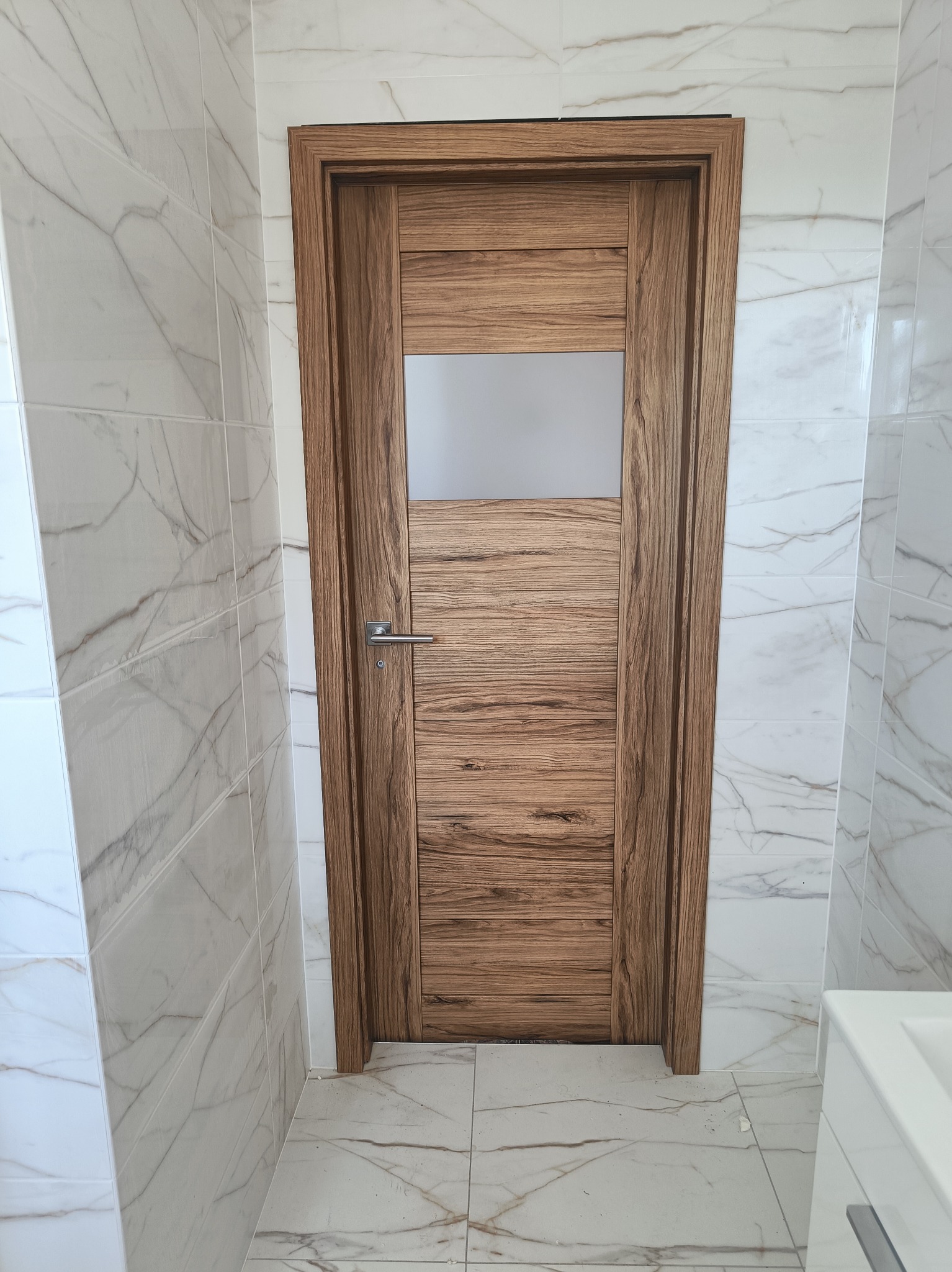 Drewniane drzwi wewnętrzne z szybą w górnej części, zamontowane w łazience z marmurowymi płytkami.