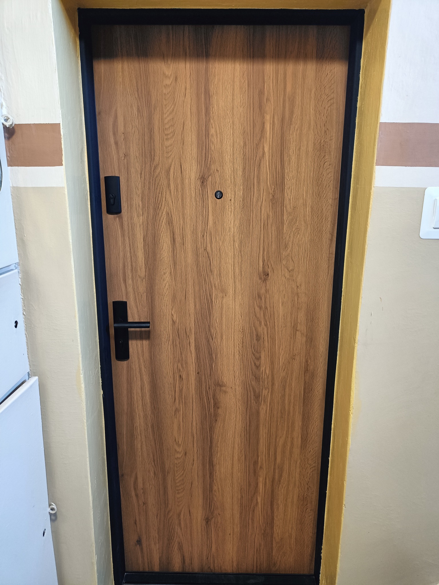 Zamknięte drzwi z drewnopodobną okleiną, czarną ościeżnicą i klamką, widoczne na tle ściany klatki schodowej z niedokładnym malowaniem.