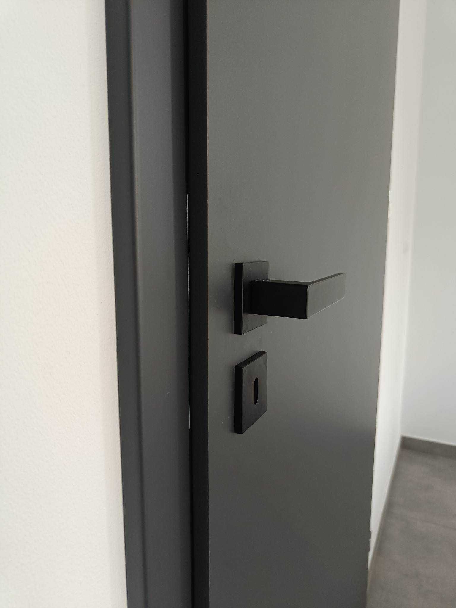 Grafitowe skrzydło drzwiowe z czarną, kwadratową klamką i minimalistycznym zamkiem na tle białej ściany i szarej podłogi.