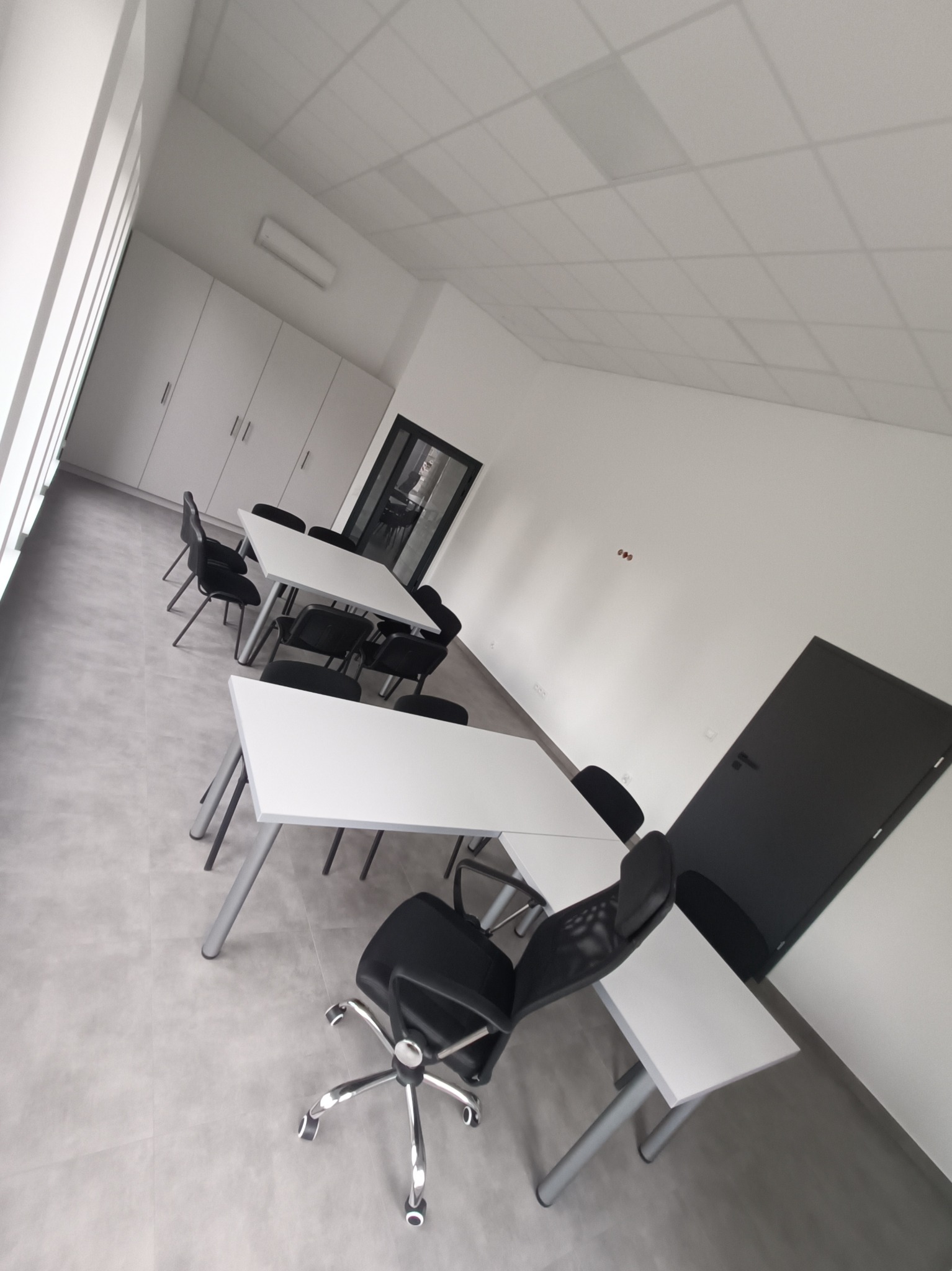 Jasne biuro z białymi biurkami i czarnymi krzesłami, widok na czarne drzwi w tle, minimalistyczny design.