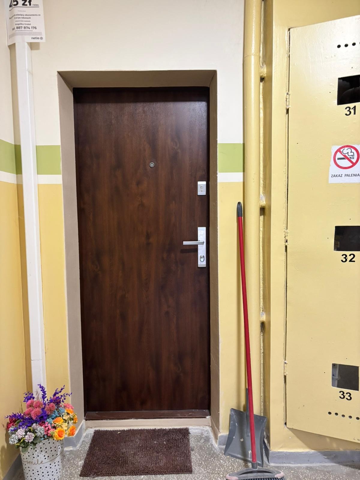 Ciemnobrązowe drzwi wejściowe do mieszkania w Strzelcach Opolskich, z wizjerem i srebrną klamką, na tle żółtej ściany z numerami skrytek pocztowych.