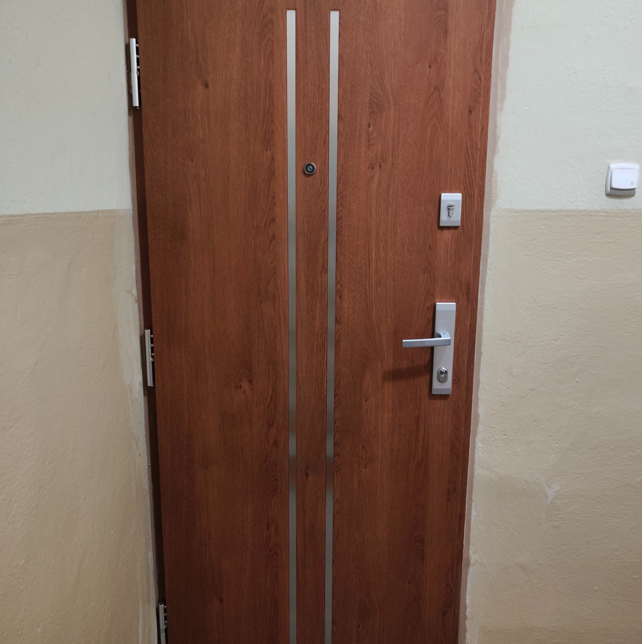 Ciemnobrązowe drzwi z dwoma srebrnymi paskami, srebrną klamką i wizjerem, zamontowane w ścianie z widocznym przełącznikiem światła.