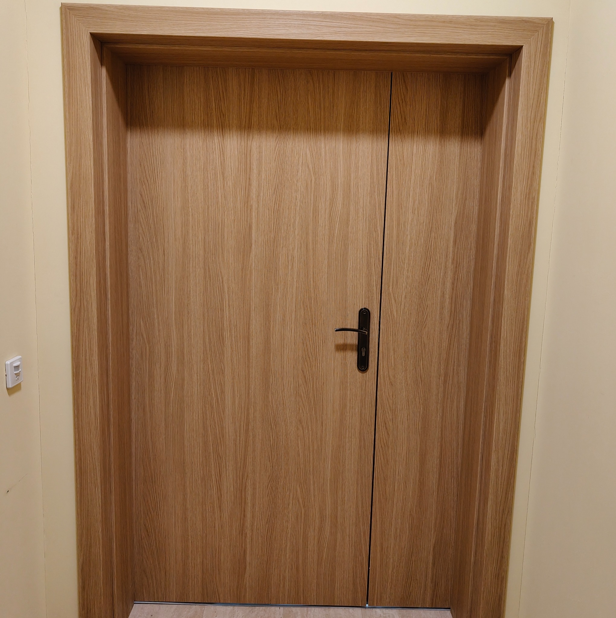 Zamknięte, drewniane drzwi wewnętrzne z czarną klamką, w jasnej ościeżnicy. Widoczny fragment ściany z włącznikiem. Minimalistyczny design.