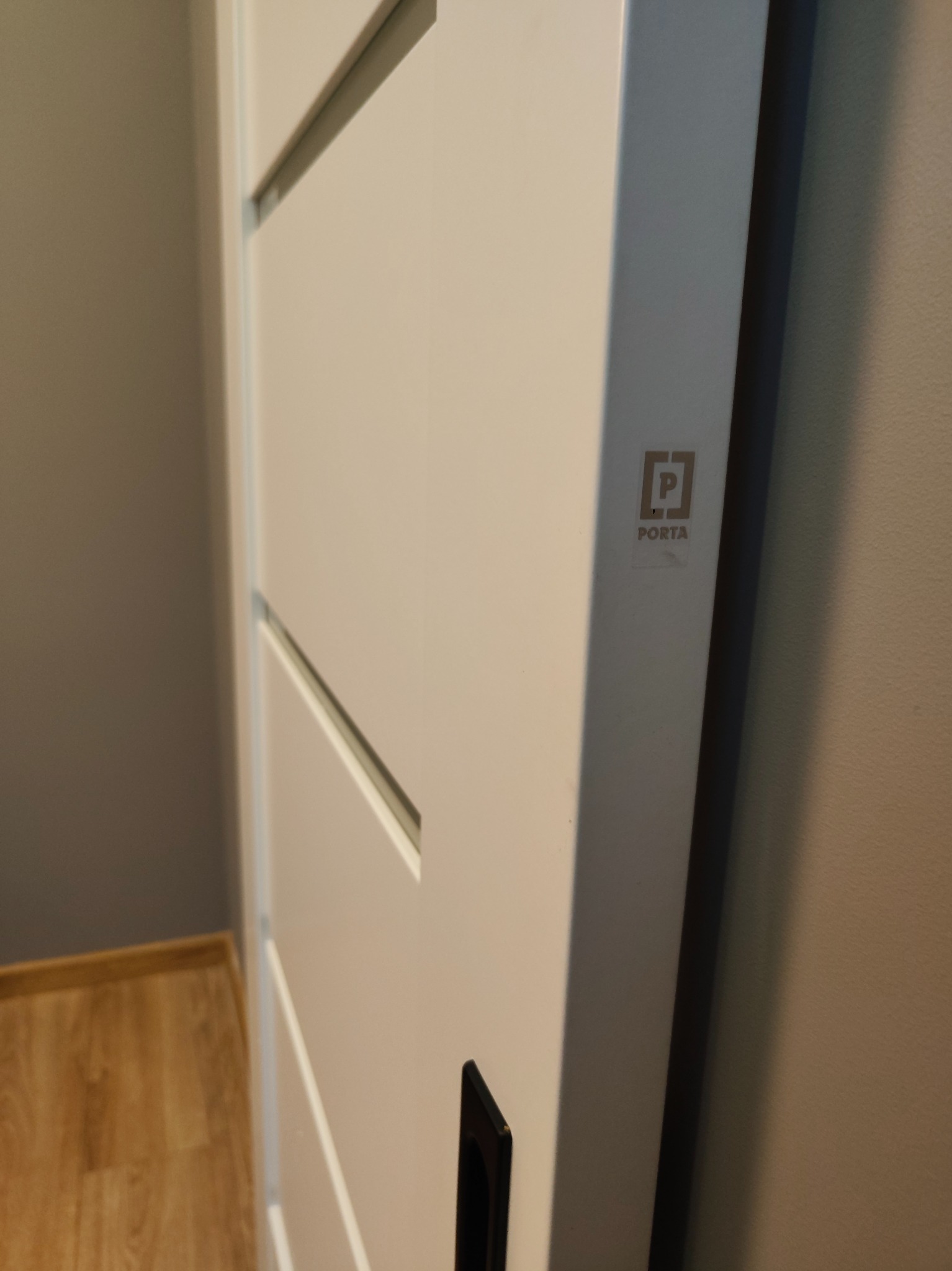 Białe drzwi wewnętrzne z poziomymi frezami i czarnym uchwytem, widoczne z boku, z logo producenta Porta na skrzydle.