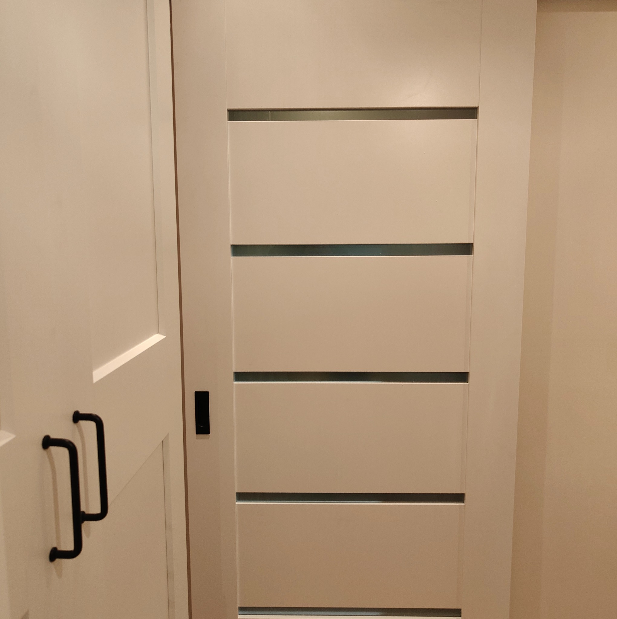 Białe drzwi wewnętrzne z czterema poziomymi przeszkleniami, widoczne czarne, metalowe okucia. Minimalistyczny design, ciepłe oświetlenie.