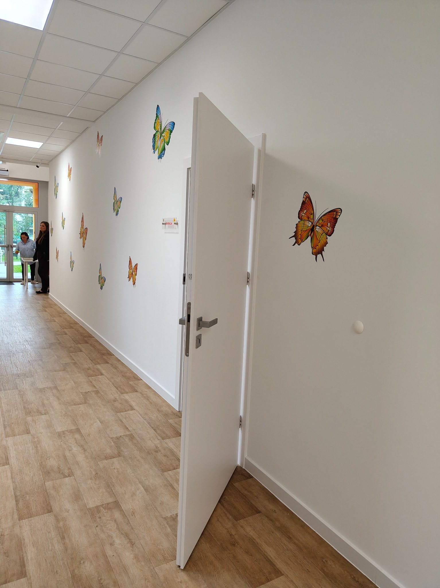 Otwarta biała stolarka drzwiowa z srebrną klamką w korytarzu z dekoracjami motyli na ścianach i panelami podłogowymi. Widoczne osoby w tle.