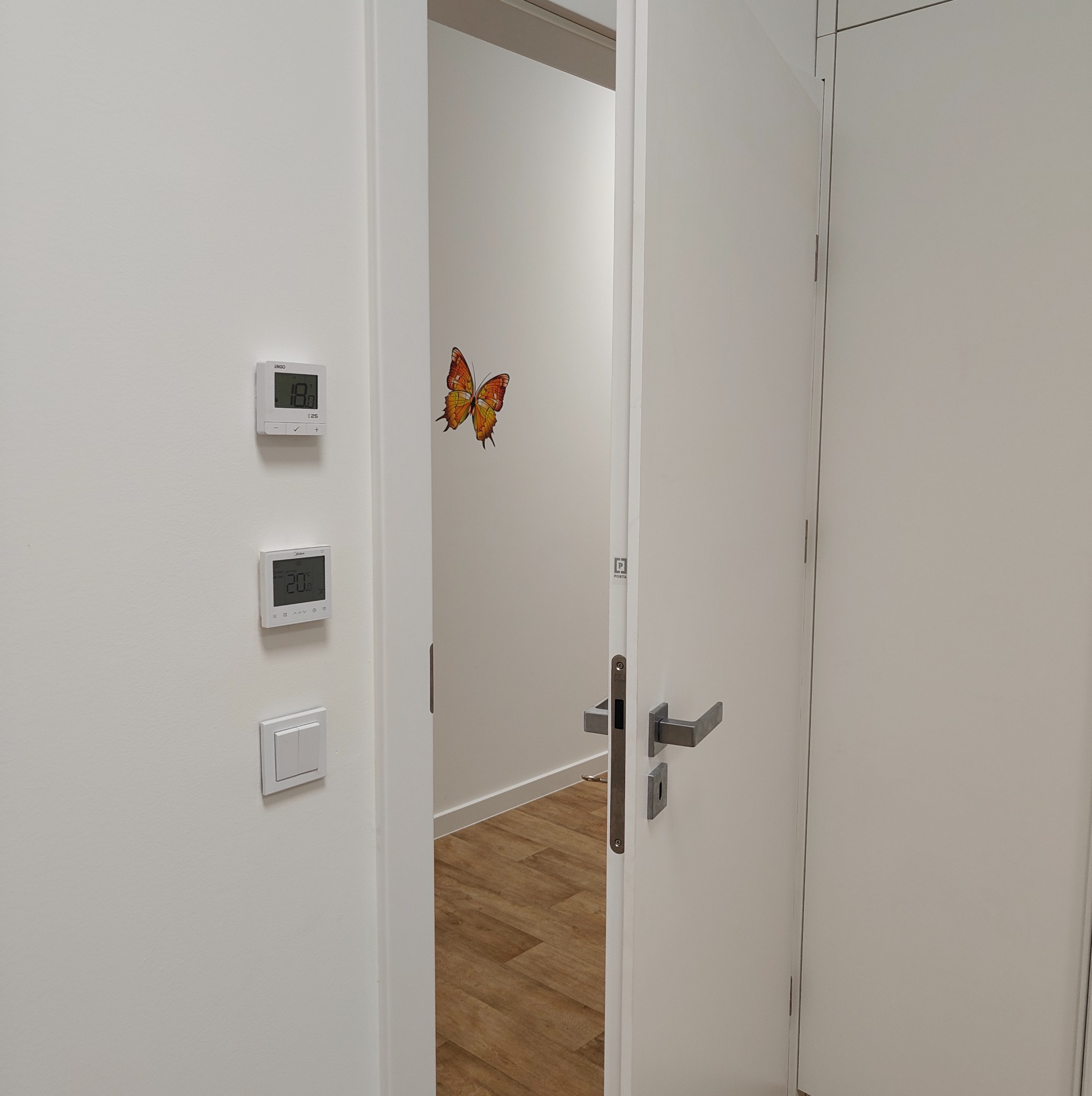 Otwarta, biała  konstrukcja z klamką, widok na wnętrze z podłogą imitującą drewno i naklejką motyla. Na ścianie termostaty i włącznik światła.