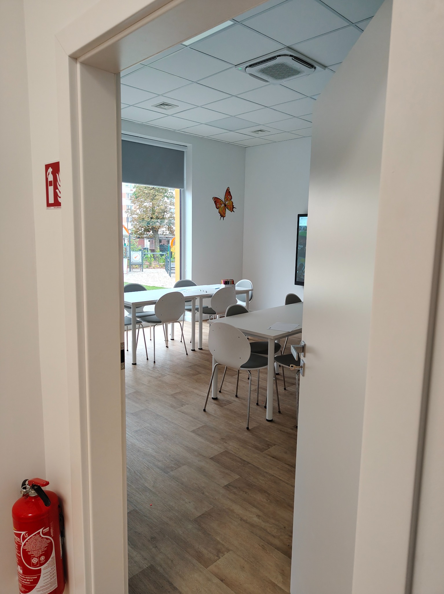 Białe drzwi uchylne, widok na jasne wnętrze z parkietem, stołami i krzesłami. Na ścianie gaśnica i dekoracja motyla. Za oknem plac zabaw.