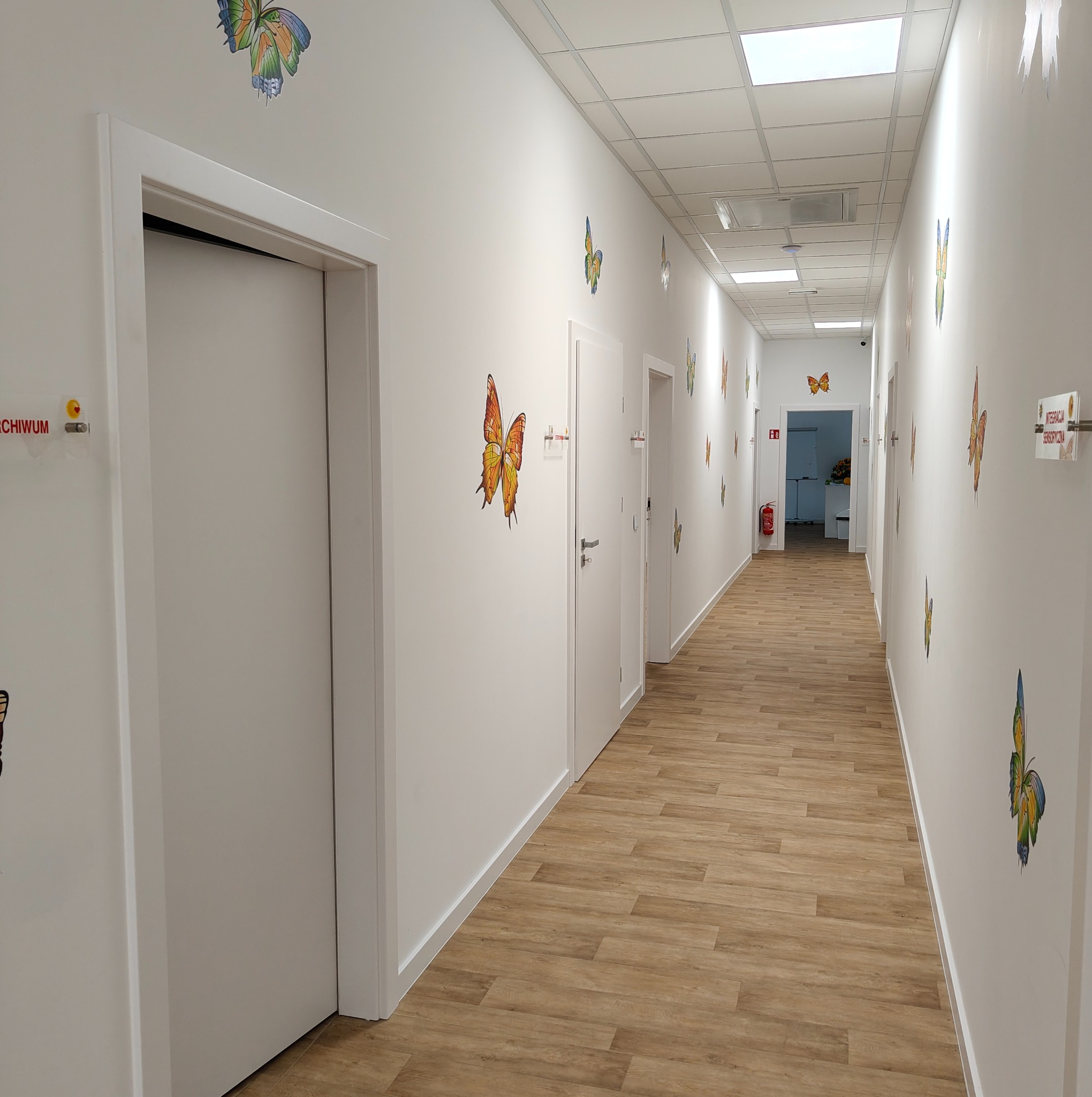 Długi korytarz z białymi drzwiami wewnętrznymi, ozdobiony naklejkami motyli. Podłoga wyłożona panelami imitującymi drewno. Widok w głąb korytarza.