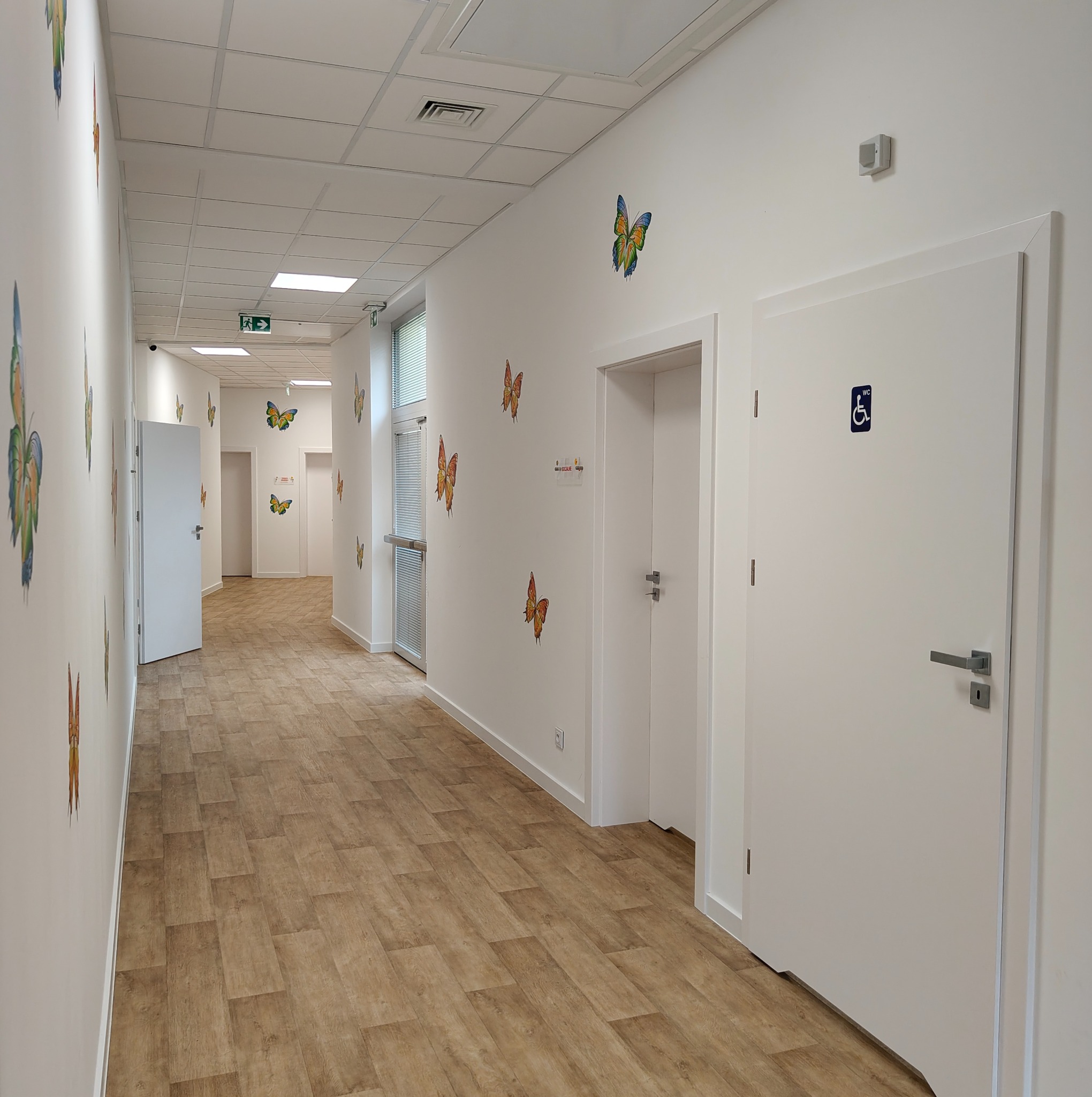 Długi korytarz w placówce medycznej z białymi drzwiami, dekoracjami motyli na ścianach i podłogą imitującą drewno. Jedne z drzwi oznaczone symbolem WC dla niepełnosprawnych.