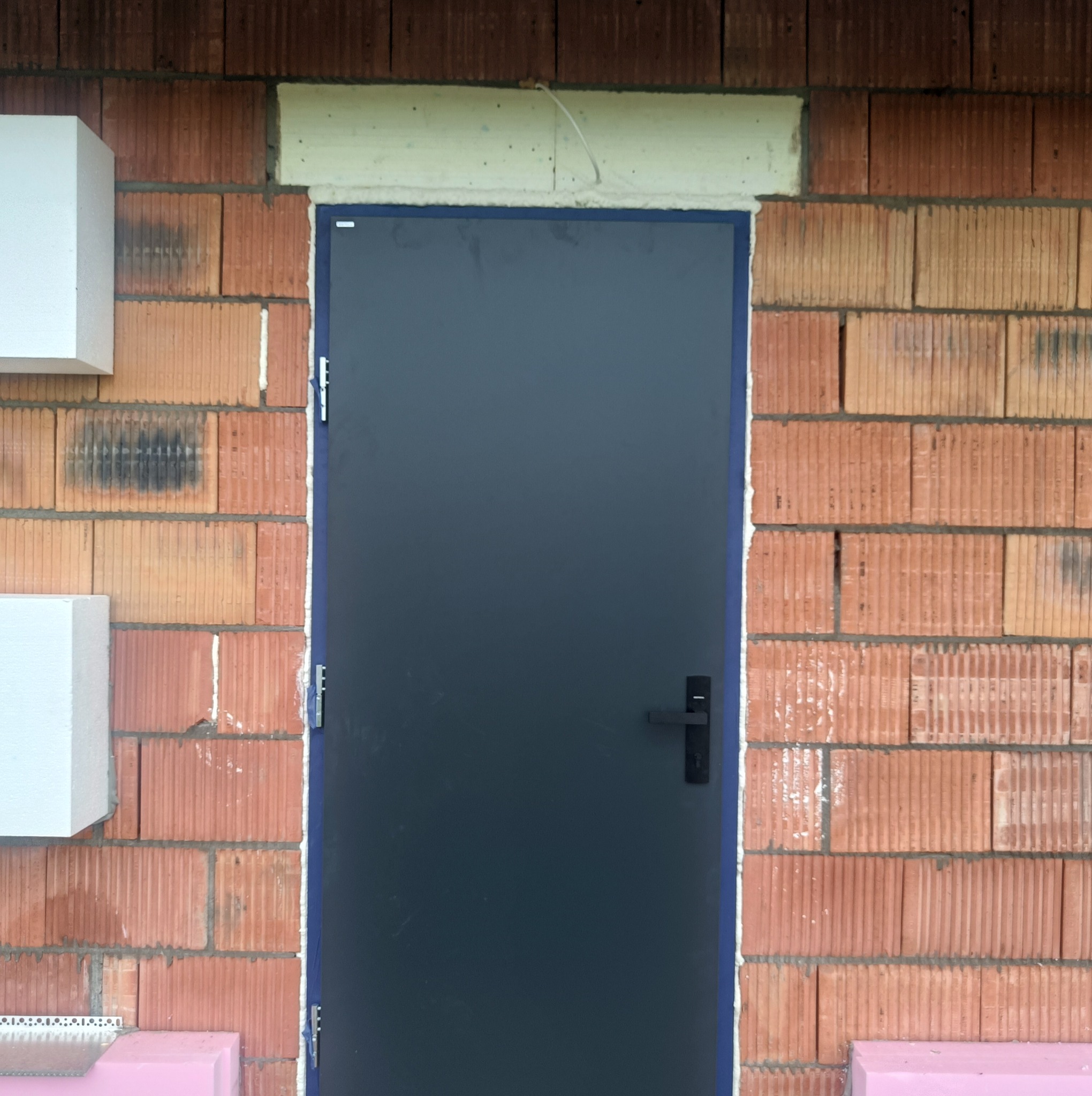 Ciemne drzwi z czarną klamką, osadzone w surowej, ceglanej ścianie z widocznym ociepleniem. Minimalistyczny design w trakcie montażu.