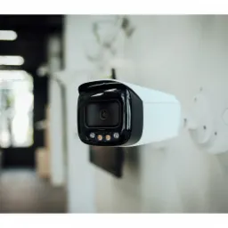 Biała kamera monitoringu z czarnym obiektywem i trzema diodami LED, zamontowana na białej ścianie, widok z boku, w tle rozmyte wnętrze pomieszczenia.