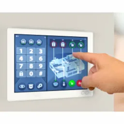 Ekran dotykowy z interfejsem graficznym przedstawiającym plan domu i panelem numerycznym, obsługiwany palcem, prawdopodobnie system sterowania bezpieczeństwem lub inteligentnym domem.