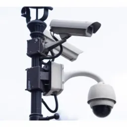 Trzy kamery przemysłowe różnego typu zamontowane na czarnym, ozdobnym słupie oświetleniowym z widocznymi przewodami i puszkami montażowymi na tle jasnego nieba.