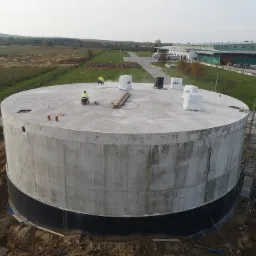 Wznoszenie betonowego, cylindrycznego zbiornika na fundamencie z izolacją, dwóch robotników na górze, widok z góry na plac budowy i okoliczne pola.