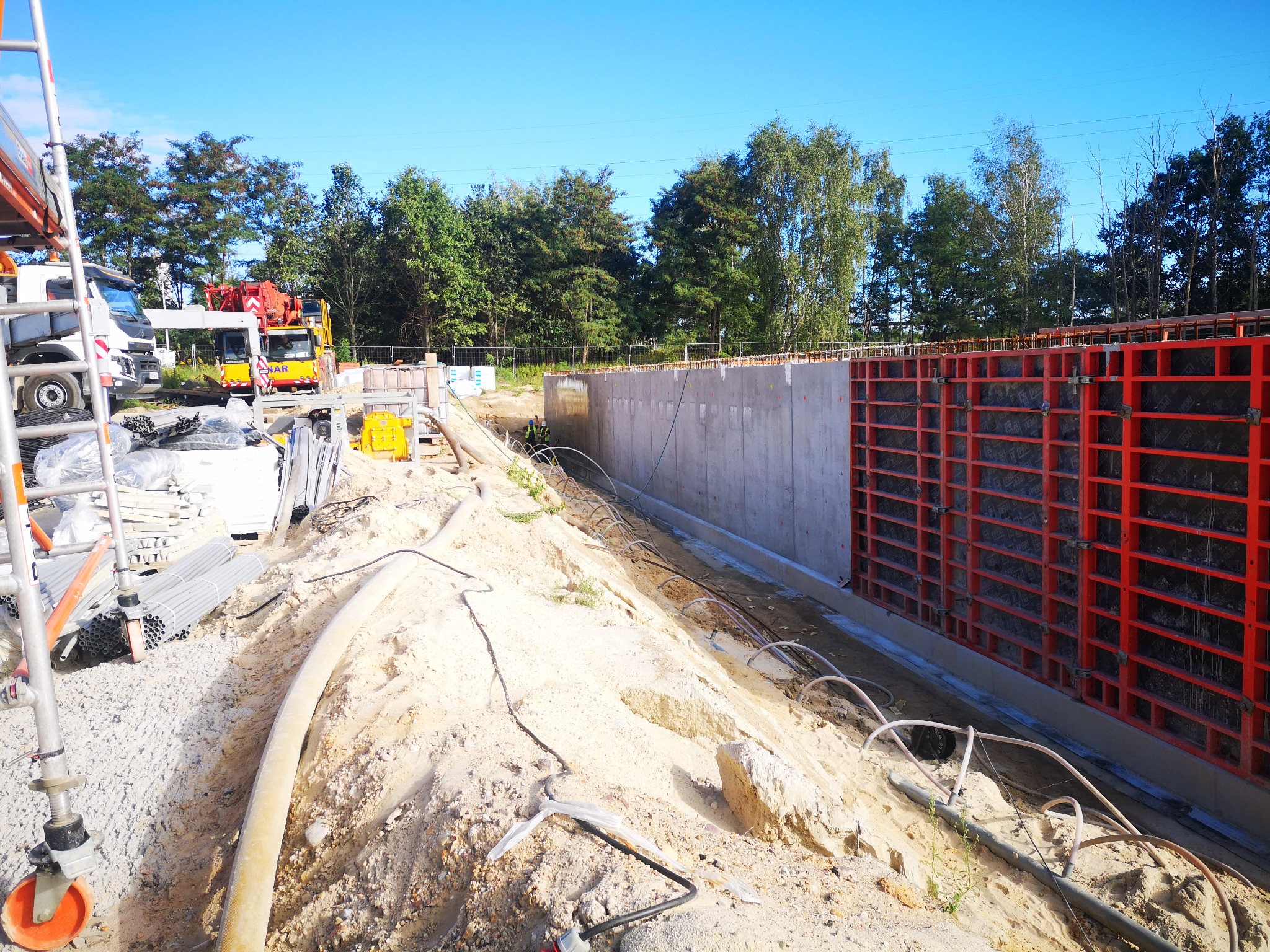 Wykop pod budowę z widocznymi elementami szalunku i zbrojenia betonowego, przygotowany do wylania betonu. W tle widoczny krajobraz z drzewami i błękitnym niebem oraz sprzęt budowlany.