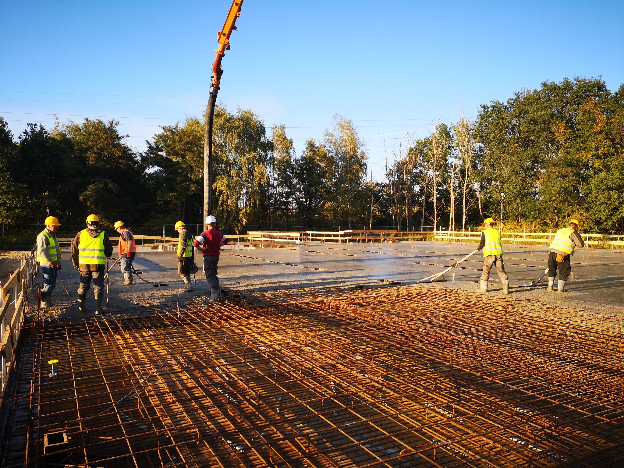 Wylewanie betonu na zbrojenie fundamentów, widoczni pracownicy w kaskach i kamizelkach odblaskowych, używana pompa do betonu, teren budowy w słoneczny dzień.