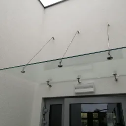 Szklany daszek nad wejściem wsparty na metalowych wspornikach, widok z dołu na szarą ramę drzwi i białą elewację budynku.