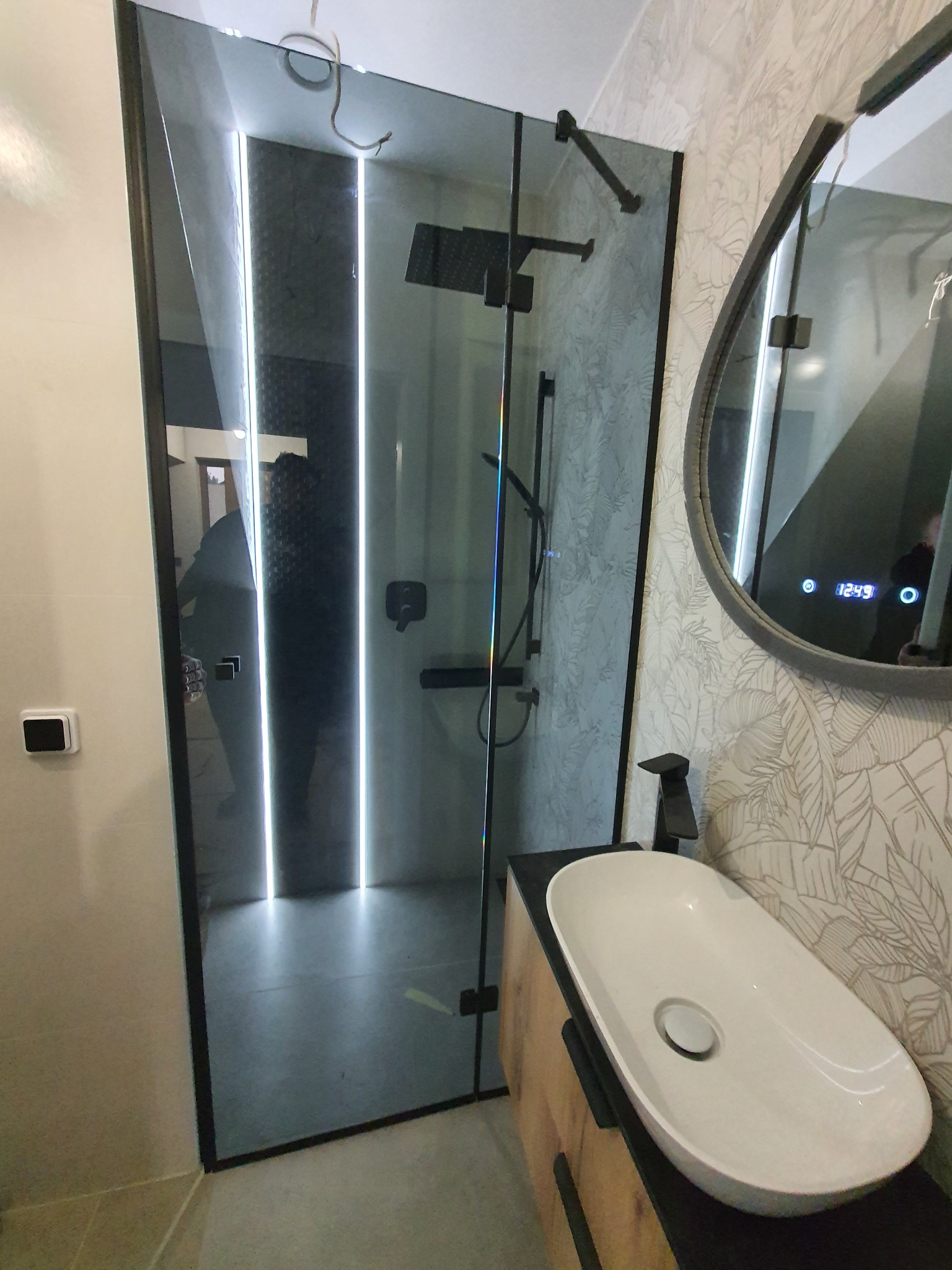 Nowoczesna kabina prysznicowa ze szklaną ścianą, czarnymi okuciami i oświetleniem LED, widoczna umywalka nablatowa i lustro z wyświetlaczem.