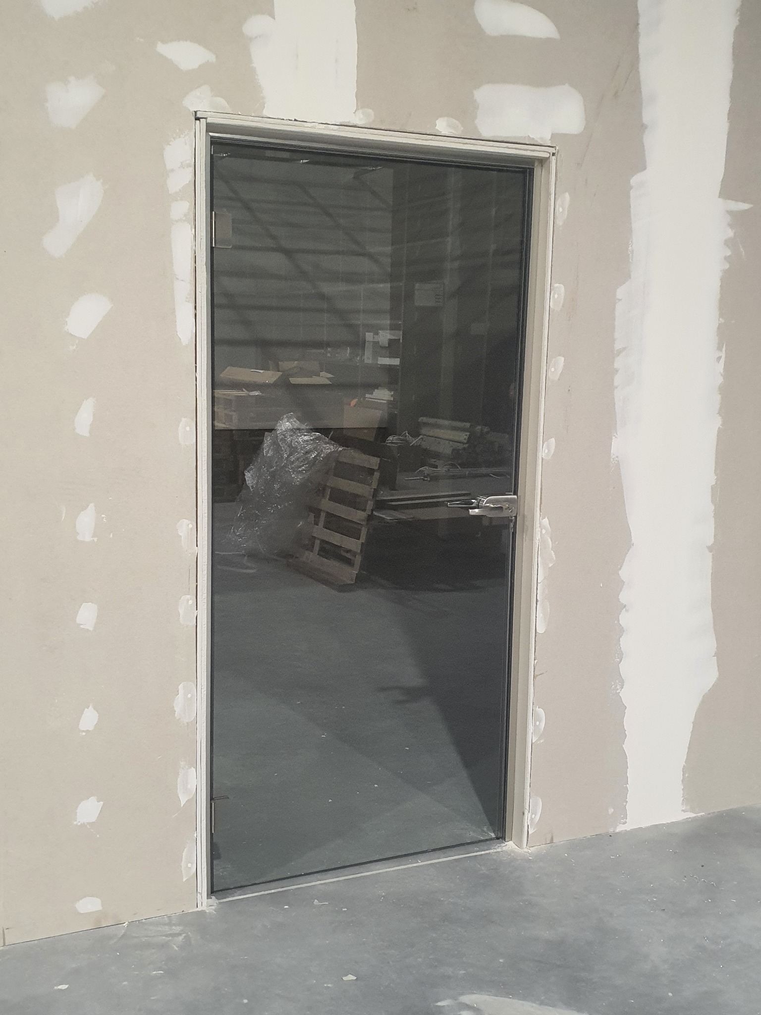 Szklane drzwi z metalową ramą i klamką, zamontowane w ścianie w trakcie wykończenia, widoczne ślady gipsu i szpachli na ścianie, w tle magazyn z paletami i zapakowanymi towarami.