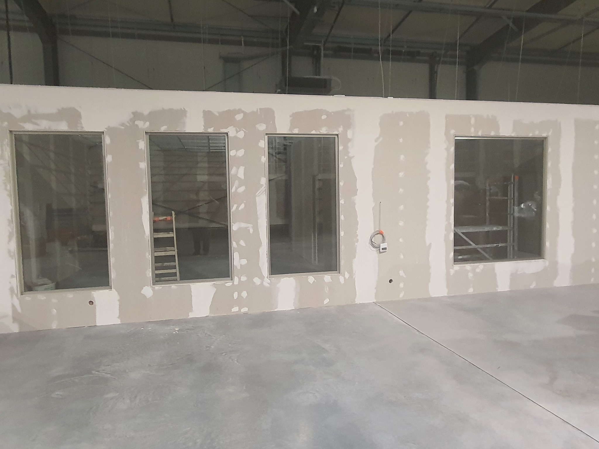 Wnętrze w trakcie wykańczania, ściana z czterema oknami w trakcie szpachlowania, widoczny szkielet konstrukcji budynku i drabina.