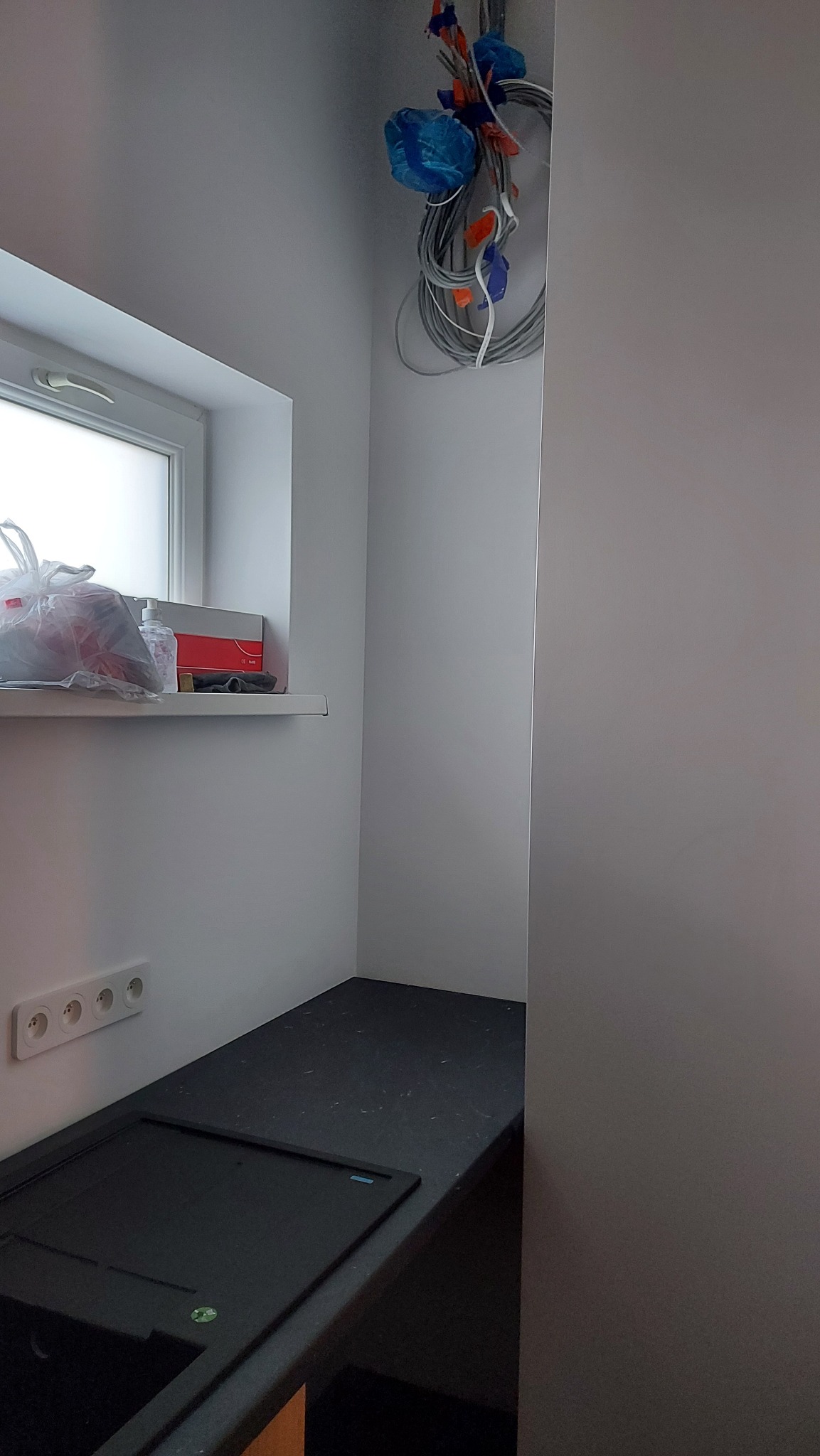 Niedokończona instalacja elektryczna w nowym budynku w Poznaniu: zwisające kable zabezpieczone niebieską folią, puszki elektryczne, okno z parapetem, gniazdka elektryczne i czarny blat.