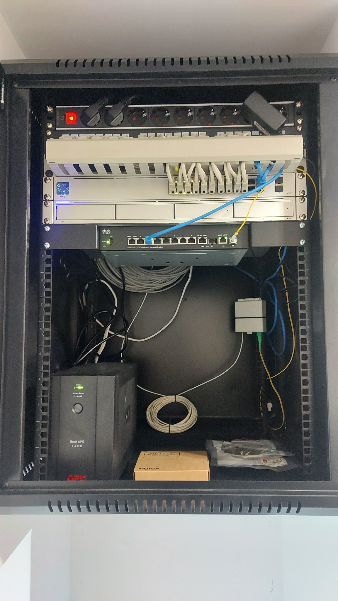 Szafa RACK z zamontowanym sprzętem sieciowym: patch panel, router Cisco, UPS APC, okablowanie strukturalne. Widoczny kabel światłowodowy i pudełko po urządzeniu Nokia.
