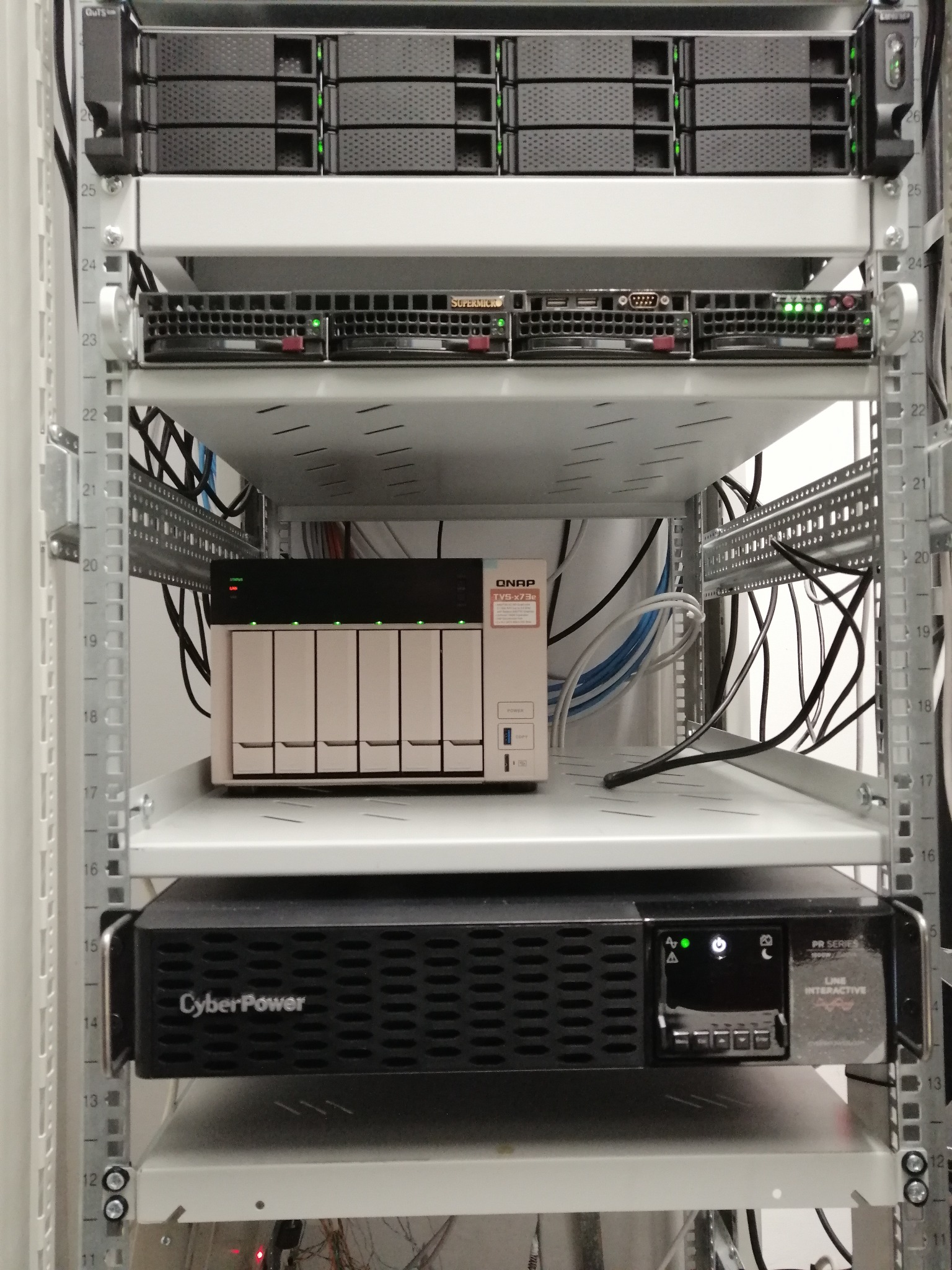 Szafa RACK z zainstalowanymi urządzeniami sieciowymi: serwerami, macierzą dyskową QNAP TVS-x73e i zasilaczem awaryjnym CyberPower PR Series, widoczne kable połączeniowe.