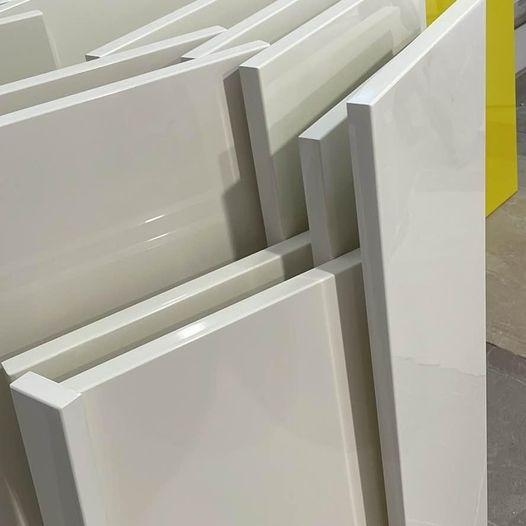 Stos białych, lakierowanych frontów meblowych z widocznym profilem krawędzi, przygotowanych do montażu, z żółtym elementem w tle.