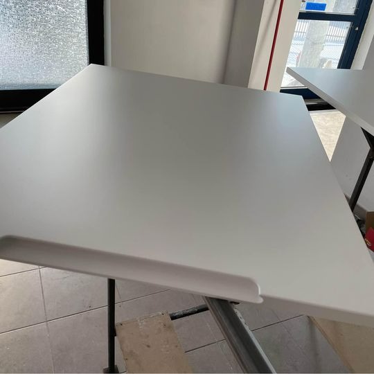 Biały, matowy blat drewniany na metalowym stelażu, widoczny fragment drugiego blatu w tle, surowe wnętrze z oknem.