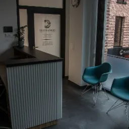 Nowoczesne wnętrze recepcji kancelarii prawnej z minimalistycznym biurkiem, dwoma turkusowymi krzesłami i widokiem na budynek z cegły za oknem.