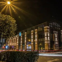 Nocny widok na nowoczesny budynek biurowy z cegły z dużymi oknami, oświetlony lampami i światłami przejeżdżających samochodów, widoczny z ulicy.