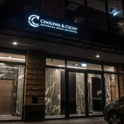 Fasada budynku kancelarii prawnej z logo 'Cholewa & Cichy' podświetlonym na czarnym tle, widoczne wejście z marmurowymi panelami i fragment zaparkowanego samochodu.