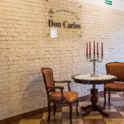 Eleganckie wnętrze restauracji z ceglaną ścianą, stolikiem z koronkową serwetą, świecznikiem z czerwonymi świecami i dwoma krzesłami w stylu vintage. Na ścianie tabliczka z nazwą restauracji Don...