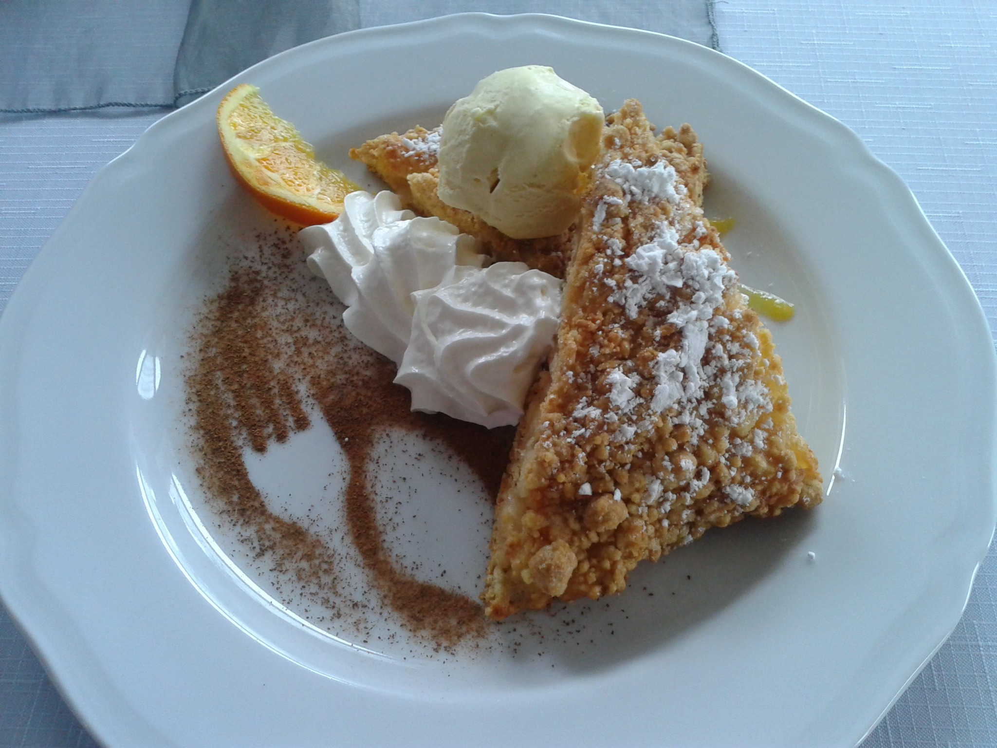 Kawałek szarlotki posypany cukrem pudrem, z bitą śmietaną i gałką lodów na białym talerzu, udekorowany plasterkiem pomarańczy i cynamonem ułożonym w kształt półksiężyca.