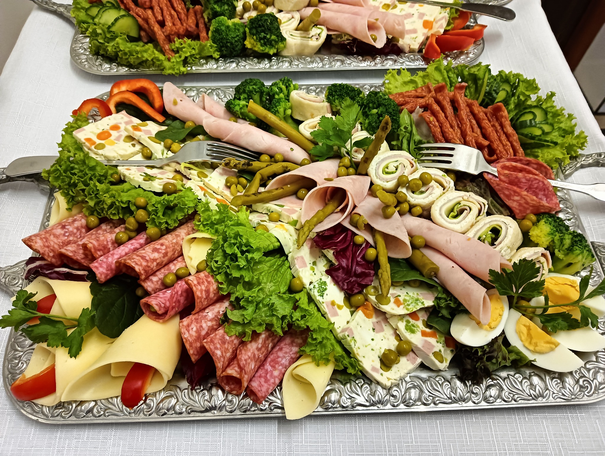 Dwa srebrne półmiski z bogatą kompozycją wędlin, serów, warzyw i jajek na białym obrusie, widoczne wędliny zwinięte w rulony, sery zawinięte z papryką, jajka na twardo, sałata, brokuły, ogórki...